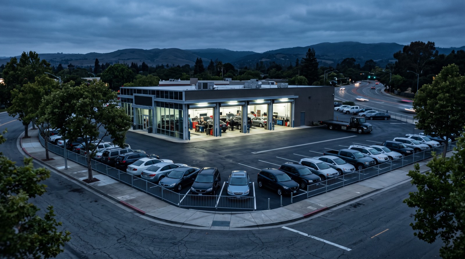 Illustrative image, not the actual property. A well kept independent automotive service and sales property on a large paved corner lot at blue hour, service bay doors open and lit, clean cars parked in rows